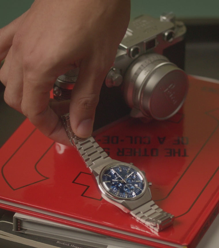 Falcon Eye Chronograph video featuring a man demonstrating putting the watch on his wrist and then laying the watch back down against a red book of Lawrence Weiner: The Other Side of A Cul-De-Sac 