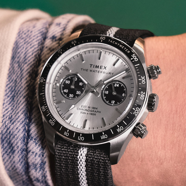 Timex Waterbury chronograph watch with a silver sunburst dial, black tachymeter bezel, two subdials, and a black fabric strap, worn on a wrist in a close-up shot.