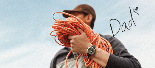 Father’s Day featuring a man outdoors holding a coil of orange climbing rope while wearing a wristwatch. The background shows a cloudy sky, with the handwritten word ‘Dad’ and a small heart graphic adding a personal, heartfelt touch.