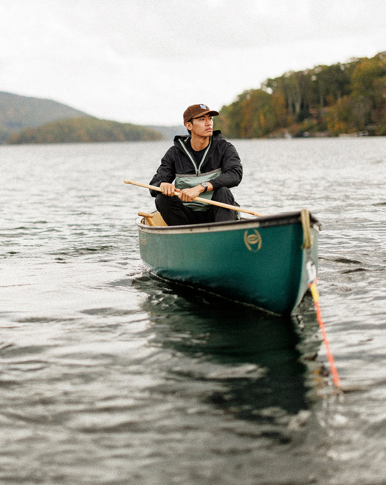 A man paddles a green canoe across a calm lake, surrounded by forested hills. He wears a brown baseball cap, black pants, and a windbreaker, and has a wristwatch on his left wrist. His expression is focused as he rows, with overcast skies and gentle ripples on the water conveying a peaceful, outdoorsy vibe.