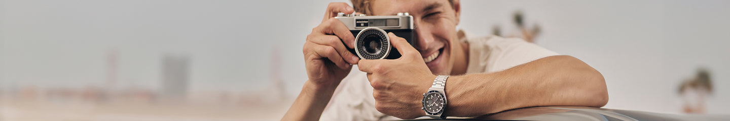 A cheerful man leans on a car while taking a photo with a vintage film camera. He wears a stainless steel chronograph watch and a white T-shirt, with a blurred beachside backdrop and palm trees in the distance.