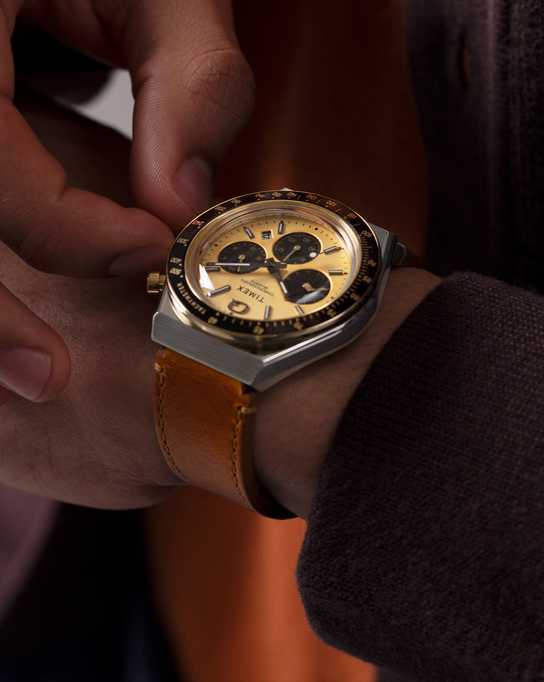 A close-up of a wrist wearing a gold and black chronograph watch with a tan leather strap. The wearer is adjusting the crown, dressed in rich-toned layers that complement the watch’s warm metallic finish.