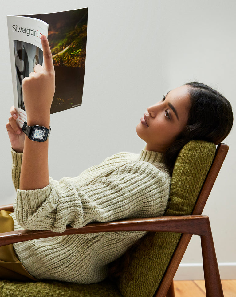 A young woman lounges comfortably on a green mid-century armchair, reading a magazine titled SilvergrainClassic. She wears a chunky knit sweater and a black digital wristwatch with a rectangular face. Her relaxed posture and serene expression suggest a quiet, contemplative moment indoors.