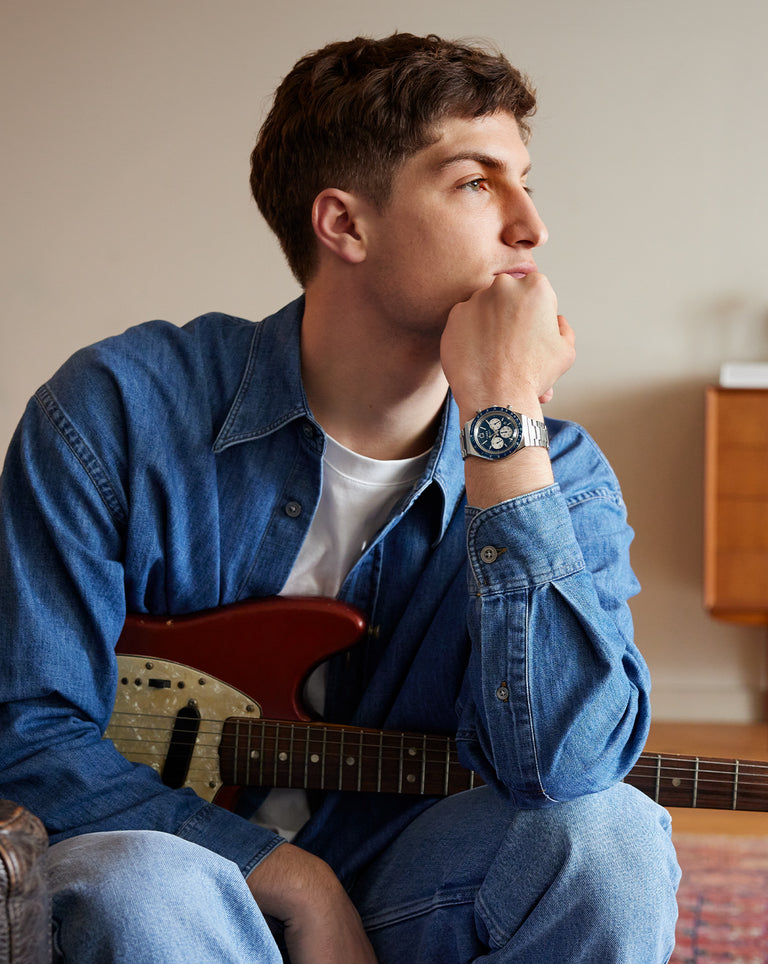 A man in a casual denim shirt and jeans sits on the floor with an electric guitar resting on his lap. He gazes thoughtfully into the distance with his chin resting on his fist. A sleek chronograph wristwatch with a blue face and metal bracelet stands out on his wrist, adding a modern edge to the laid-back setting.