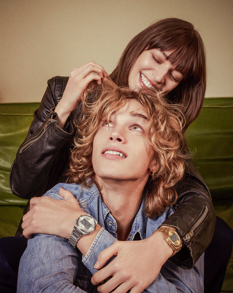 A young couple sits playfully together. The woman, smiling, wraps her arms around the man’s shoulders. Both wear retro digital watches—hers gold-toned, his silver—adding a nostalgic touch to their stylish, casual outfits.