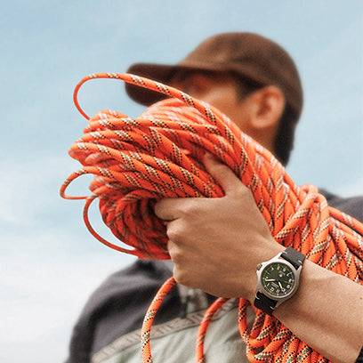 A person holding a coiled orange climbing rope, wearing a Timex Expedition watch with a green dial and black strap. The person’s face is slightly out of focus, emphasizing the rugged gear and timepiece.