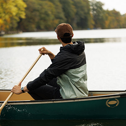 A man in a cap and two-tone jacket paddles a green canoe on a calm, tree-lined lake, wearing a Timex Expedition watch on his left wrist.