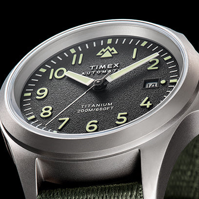 Close-up studio image of a Timex Expedition watch with a black textured dial, luminous hands and markers, date window, and titanium 200M water resistance rating, set against a dark background.