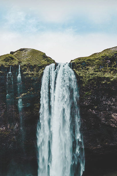 A powerful waterfall cascades between green, moss-covered cliffs under a cloudy sky, creating a dramatic and natural landscape.