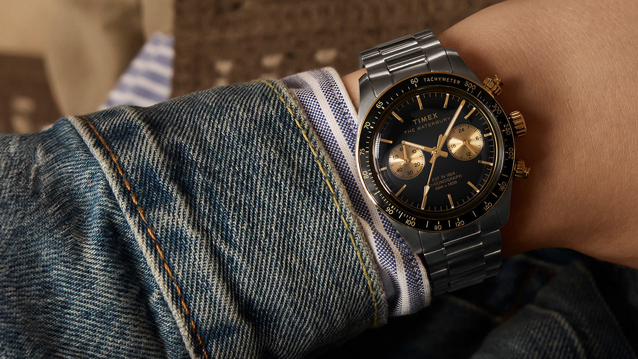 A Timex Waterbury Heritage Chronograph with a black dial and gold-tone subdials worn on the wrist, paired with a denim jacket and striped dress shirt, showcasing a bold yet classic casual look.
