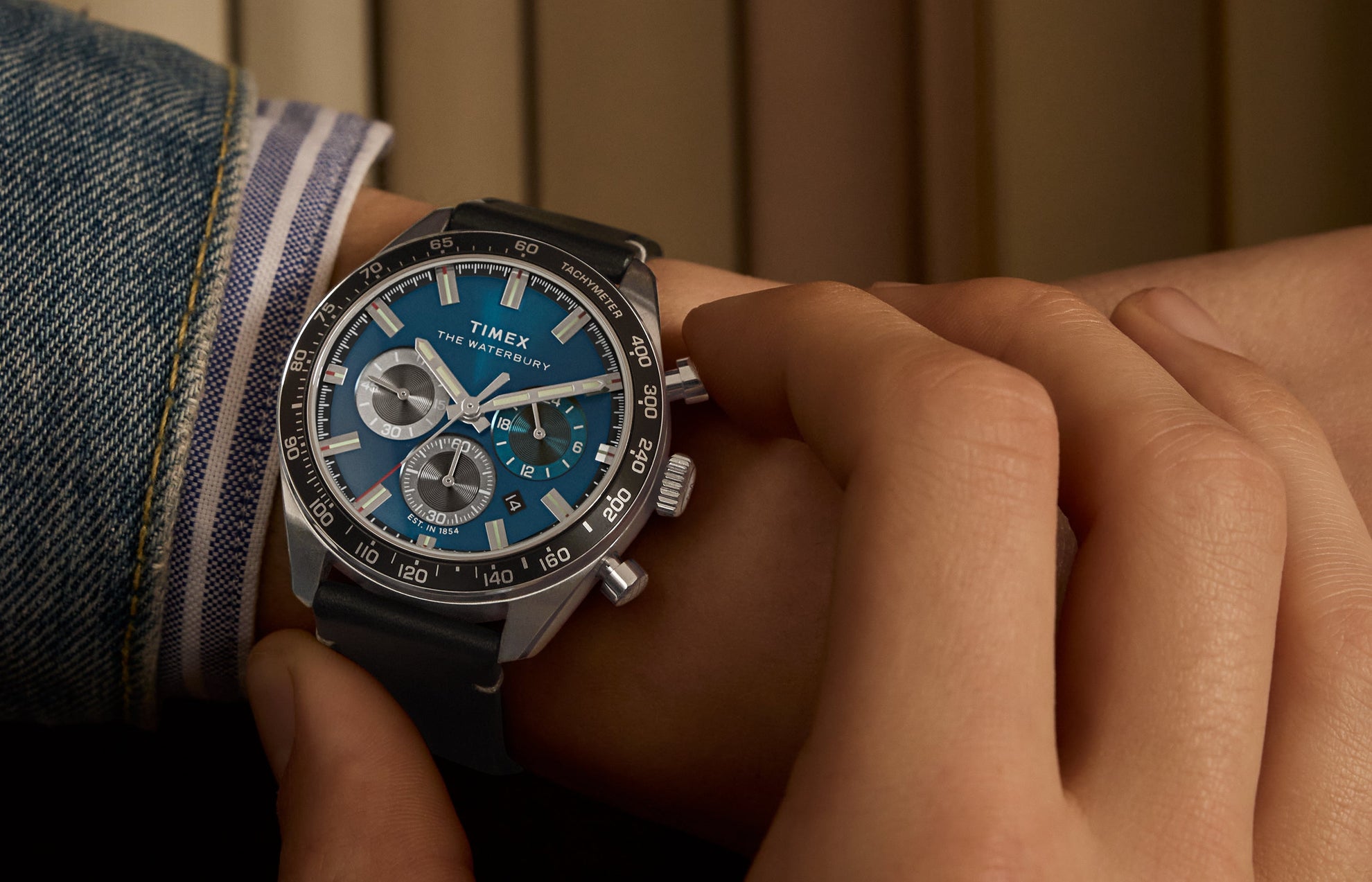 A person adjusts the crown of a Timex Waterbury Chronograph watch on their wrist. The blue dial contrasts against silver subdials and luminous markers, framed by a tachymeter bezel. The black leather strap and rolled-up denim cuff complete the casual, yet polished look.