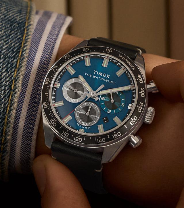 A person adjusts the crown of a Timex Waterbury Chronograph watch on their wrist. The blue dial contrasts against silver subdials and luminous markers, framed by a tachymeter bezel. The black leather strap and rolled-up denim cuff complete the casual, yet polished look.