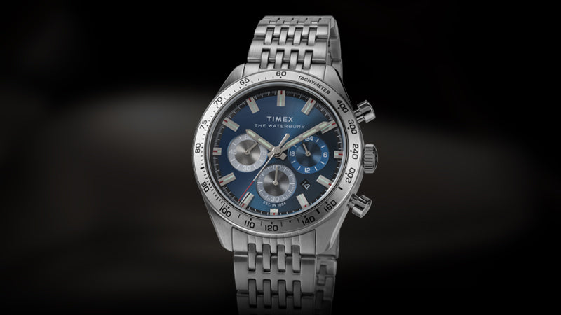 Front-facing product image of the Timex Waterbury Chronograph watch with a stainless steel bracelet and case. The deep blue dial features three subdials in silver and blue, a date window at 4 o’clock, and luminous hands and markers. The tachymeter bezel adds a sporty and functional touch to the elegant design, set against a dark gradient background.