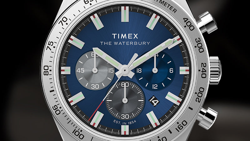 Close-up of the Timex Waterbury Chronograph dial, highlighting the blue sunburst texture and contrasting subdials. Details include luminous baton hour markers with red and green accents, a red-tipped second hand, and crisp chronograph registers. The bezel markings and subtle branding “Est. in 1854” reinforce the watch’s heritage and precision.