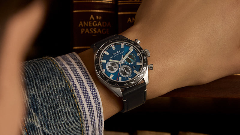 Close-up of the Timex Waterbury Chronograph on a wrist, featuring a striking blue dial, three subdials, and a tachymeter bezel. The watch is paired with a black leather strap and worn with a denim jacket and striped shirt cuff. In the background, leather-bound books—one titled “A to Anegada Passage”—add a refined, intellectual atmosphere.