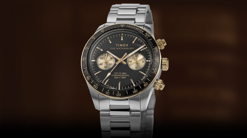 Front product view of the Timex Waterbury Heritage Chronograph featuring a black dial, gold subdials, a black tachymeter bezel, and a two-tone design with stainless steel case and bracelet.