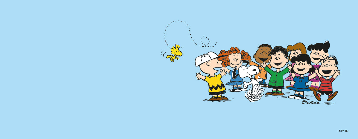 A cheerful illustration of Peanuts characters on a light blue background. Charlie Brown stands in the center wearing a yellow zigzag shirt and white cap, smiling as Woodstock the bird flies playfully above him. Snoopy joyfully jumps beside him while friends including Lucy, Linus, Peppermint Patty, Franklin, Sally, and others gather around laughing and clapping. The scene radiates warmth, friendship, and classic Peanuts charm.