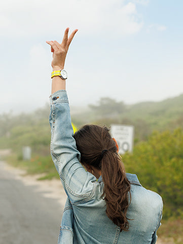 Person wearing a denim jacket and yellow watch raises their arm in a peace sign while walking down a misty country road.