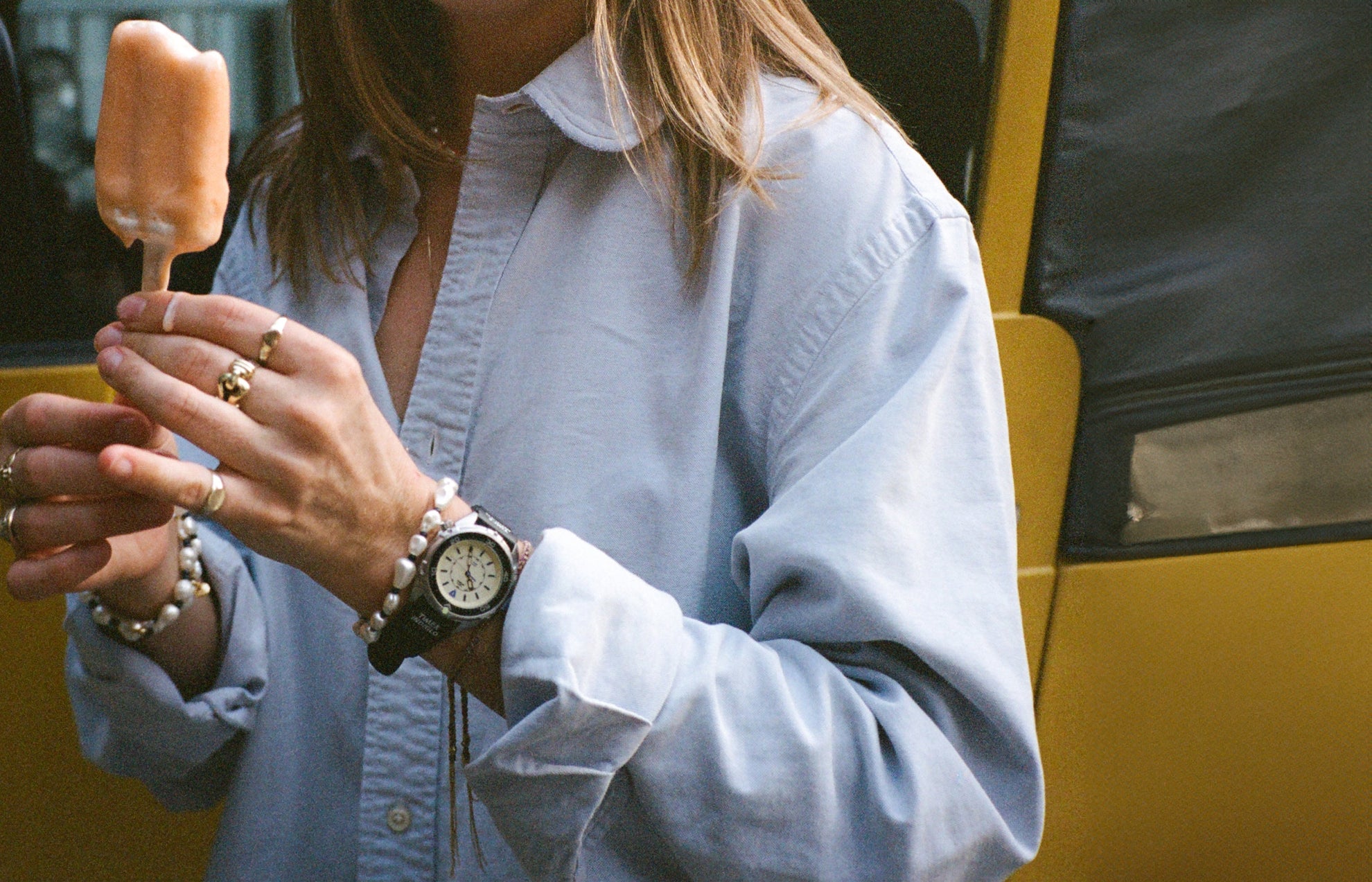 A close-up of a woman holding a partially eaten orange ice pop. She wears a light blue oversized shirt and multiple rings. Her wrist features the black Timex x Dimepiece watch with a pearlescent beaded bracelet beside it.