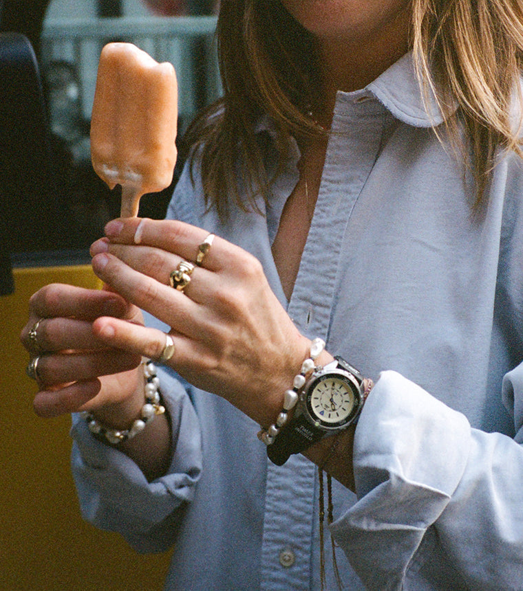 A close-up of a woman holding a partially eaten orange ice pop. She wears a light blue oversized shirt and multiple rings. Her wrist features the black Timex x Dimepiece watch with a pearlescent beaded bracelet beside it.