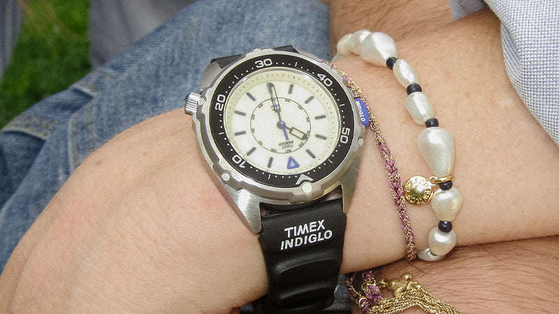 Detailed close-up of the Timex x Dimepiece watch on a person's wrist. The black rubber strap features the “TIMEX INDIGLO” logo, and the white dial includes a bold black chapter ring and triangle marker at 12 o'clock. A pearl bracelet and other fine accessories accompany the watch.