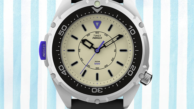 Digital render of the Timex x Dimepiece watch, showing a detailed front view. The black rotating bezel contrasts with a cream-colored dial. Bold black hands and indices frame a 60-second inner ring. The purple triangle at 12 o’clock and INDIGLO button are prominent.
