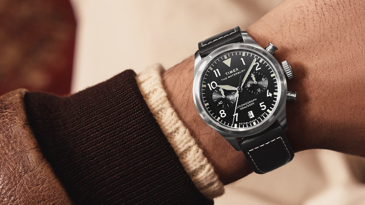 A man wearing the Timex Waterbury Ace Chronograph on his wrist, featuring a black dial with white numerals, chronograph subdials, and a black leather strap with white stitching.