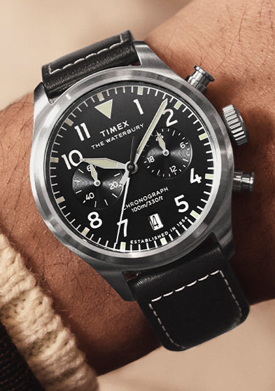 A man wearing the Timex Waterbury Ace Chronograph on his wrist, featuring a black dial with white numerals, chronograph subdials, and a black leather strap with white stitching.