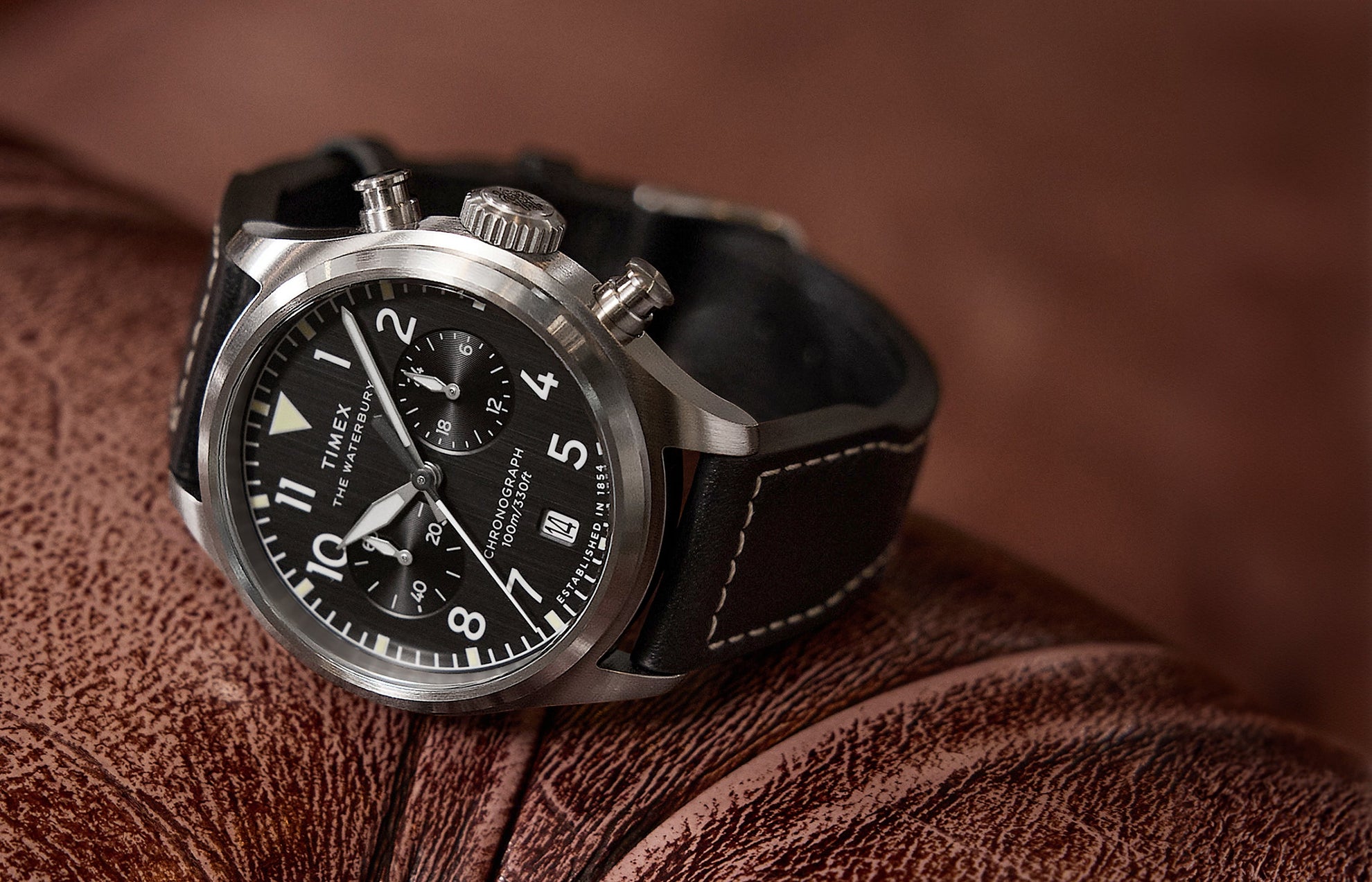 Timex Waterbury Ace Chronograph resting on a brown leather surface, showcasing its brushed stainless-steel case, black dial, chronograph subdials, and stitched leather strap.