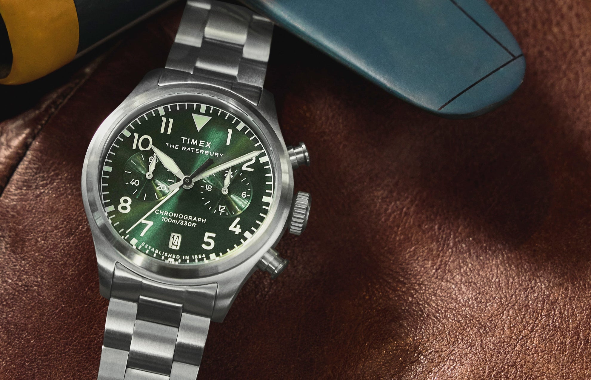 Timex Waterbury Ace Chronograph with a green dial and stainless steel bracelet resting on a brown leather surface, highlighting its brushed steel case and chronograph subdials.