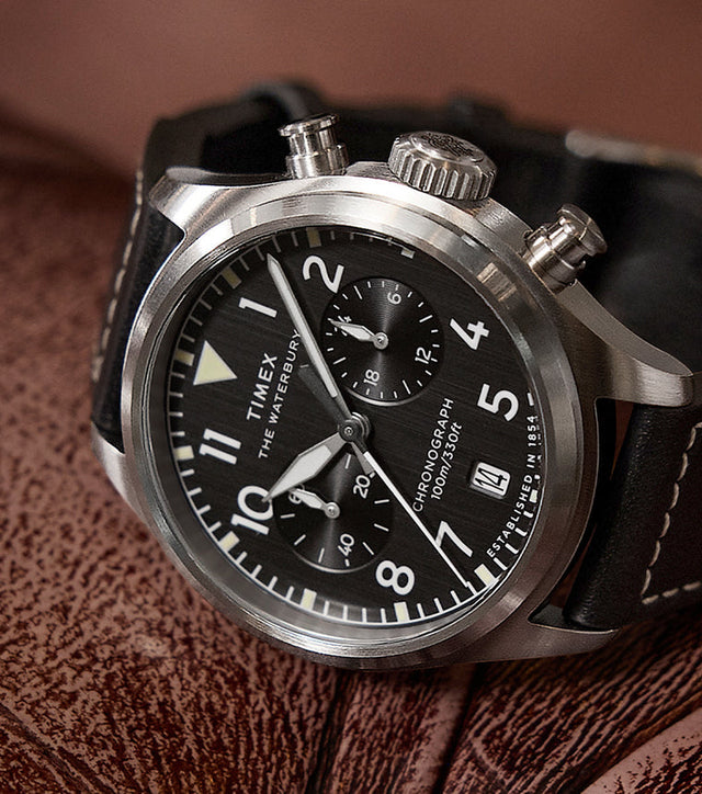 Timex Waterbury Ace Chronograph resting on a brown leather surface, showcasing its brushed stainless-steel case, black dial, chronograph subdials, and stitched leather strap.