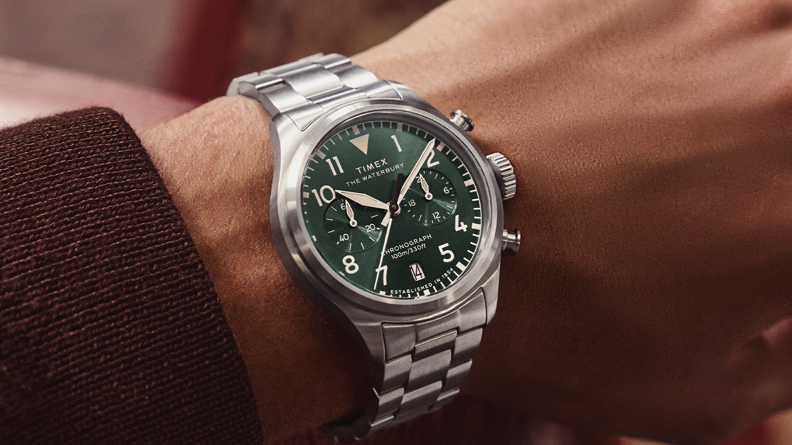 Close-up of the Timex Waterbury Ace Chronograph with a green dial and stainless steel bracelet, worn on the wrist. The watch features luminous hands, chronograph subdials, bold Arabic numerals, and a date window at 6 o’clock.