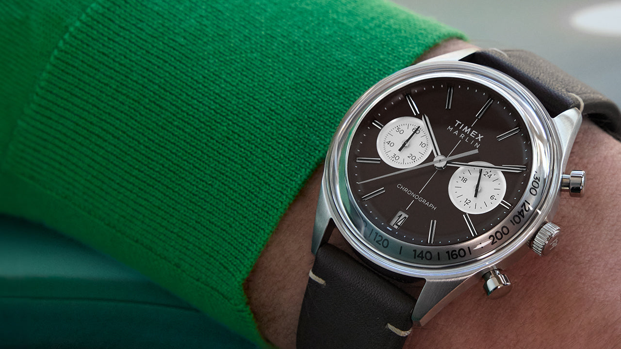 Close-up of the Timex Marlin® Jet Quartz Chronograph on wrist, featuring a rich brown dial, two contrasting white sub-dials, a date window at 6 o’clock, silver-tone case, and brown leather strap. The wearer is dressed in a green sweater.