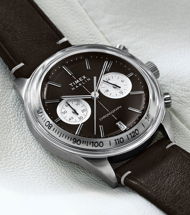 The Timex Marlin® Jet Chronograph is shown resting on a soft white backdrop. The polished case highlights the vintage-inspired brown dial and white dual sub-dials, complemented by a stitched brown leather strap.