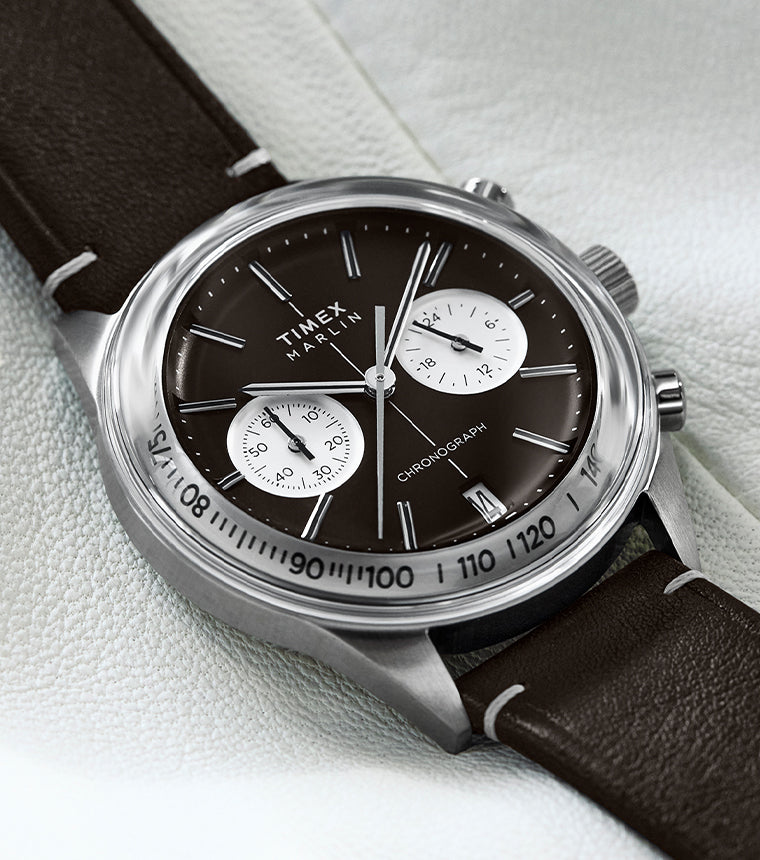 The Timex Marlin® Jet Chronograph is shown resting on a soft white backdrop. The polished case highlights the vintage-inspired brown dial and white dual sub-dials, complemented by a stitched brown leather strap.