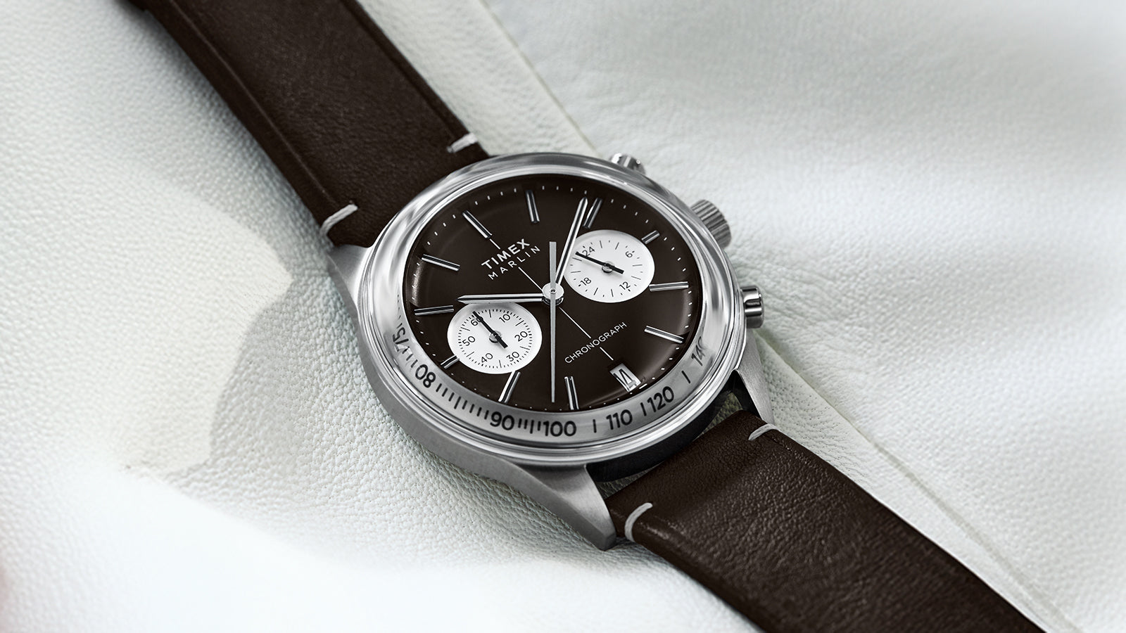 The Timex Marlin® Jet Chronograph is shown resting on a soft white backdrop. The polished case highlights the vintage-inspired brown dial and white dual sub-dials, complemented by a stitched brown leather strap.