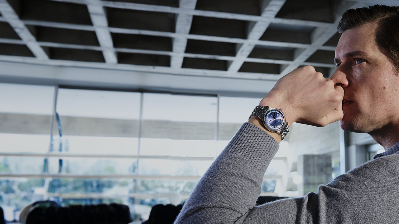A man in a gray sweater sits in a modern setting with a pensive expression, showing off the Timex Marlin® Quartz GMT watch on his wrist. The watch features a deep blue dial with a stainless steel bracelet and red arrow GMT hand.