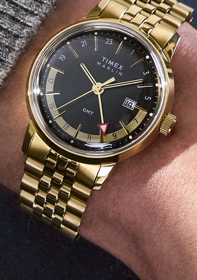 A close-up of a person wearing the Timex Marlin® Quartz GMT with a gold-tone case and bracelet. The watch features a black dial, gold-tone hands and indices, a red GMT hand, and a date display at 3 o'clock.