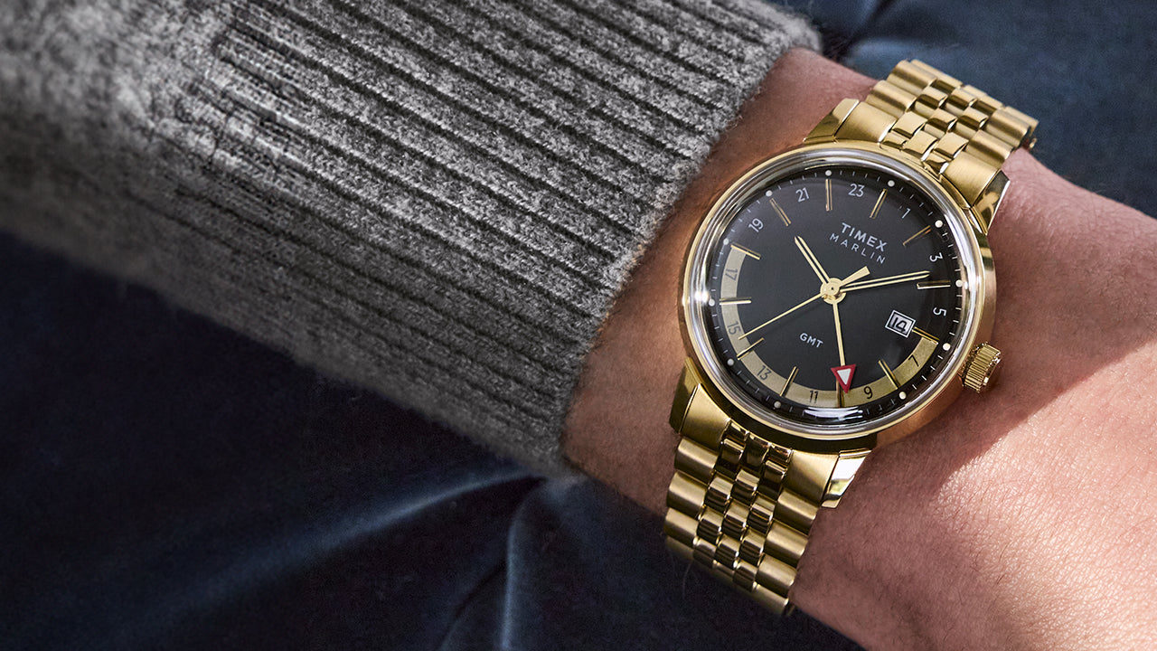 A close-up of a person wearing the Timex Marlin® Quartz GMT with a gold-tone case and bracelet. The watch features a black dial, gold-tone hands and indices, a red GMT hand, and a date display at 3 o'clock.