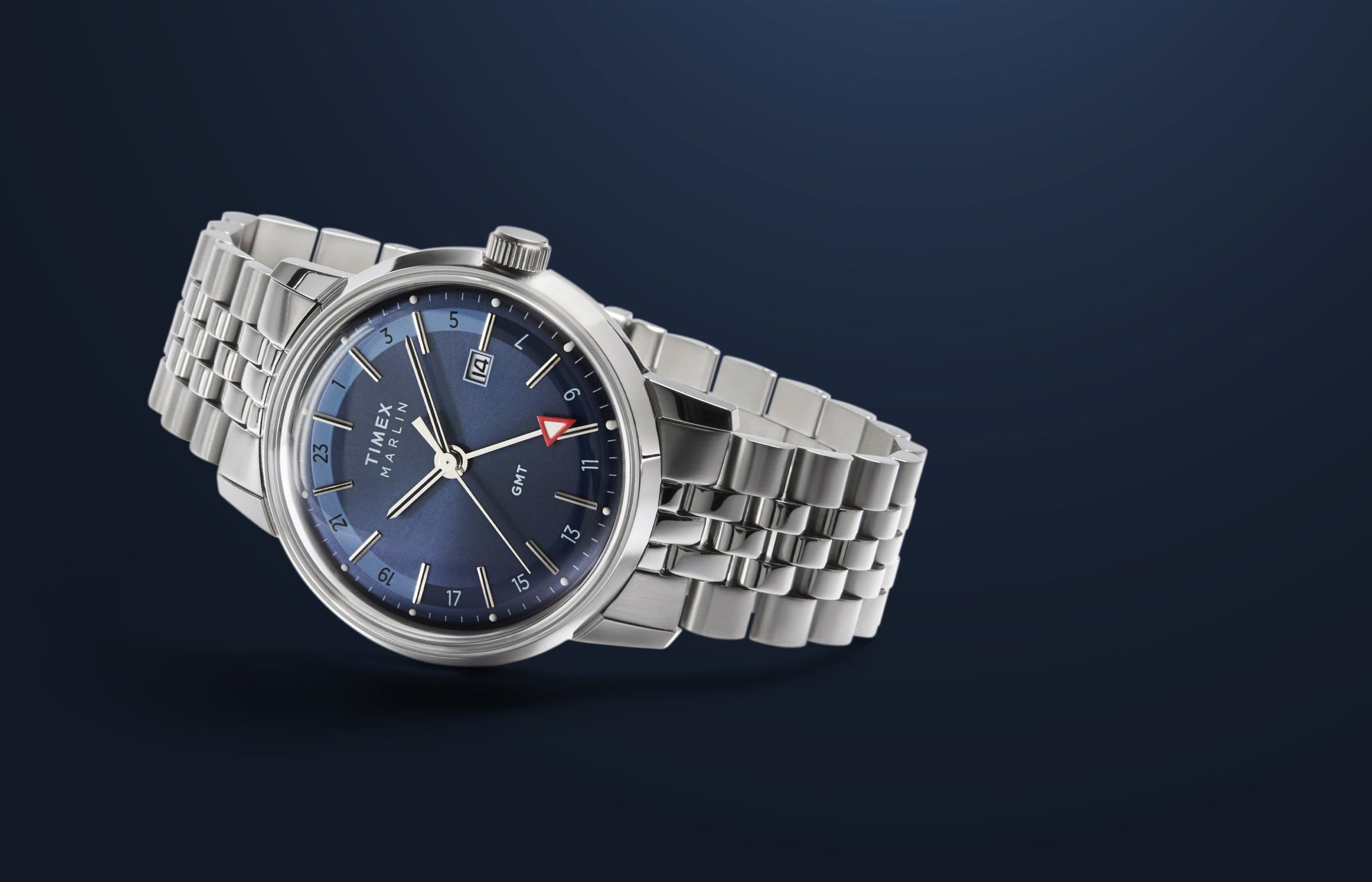 A Timex Marlin® Quartz GMT wristwatch with a deep blue sunray dial, silver-tone hour markers, a date display at 3 o’clock, and a red-arrow GMT hand. The watch has a polished stainless-steel case and a multi-link stainless-steel bracelet, shown against a dark blue gradient background.