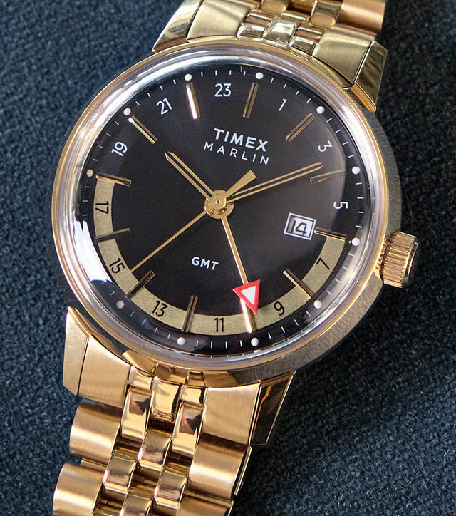 The Timex Marlin® Quartz GMT in gold-tone rests on a dark textured surface. Its black dial contrasts with golden accents, and the red arrow GMT hand adds a pop of color. The bracelet and case shine in a polished gold-tone finish.