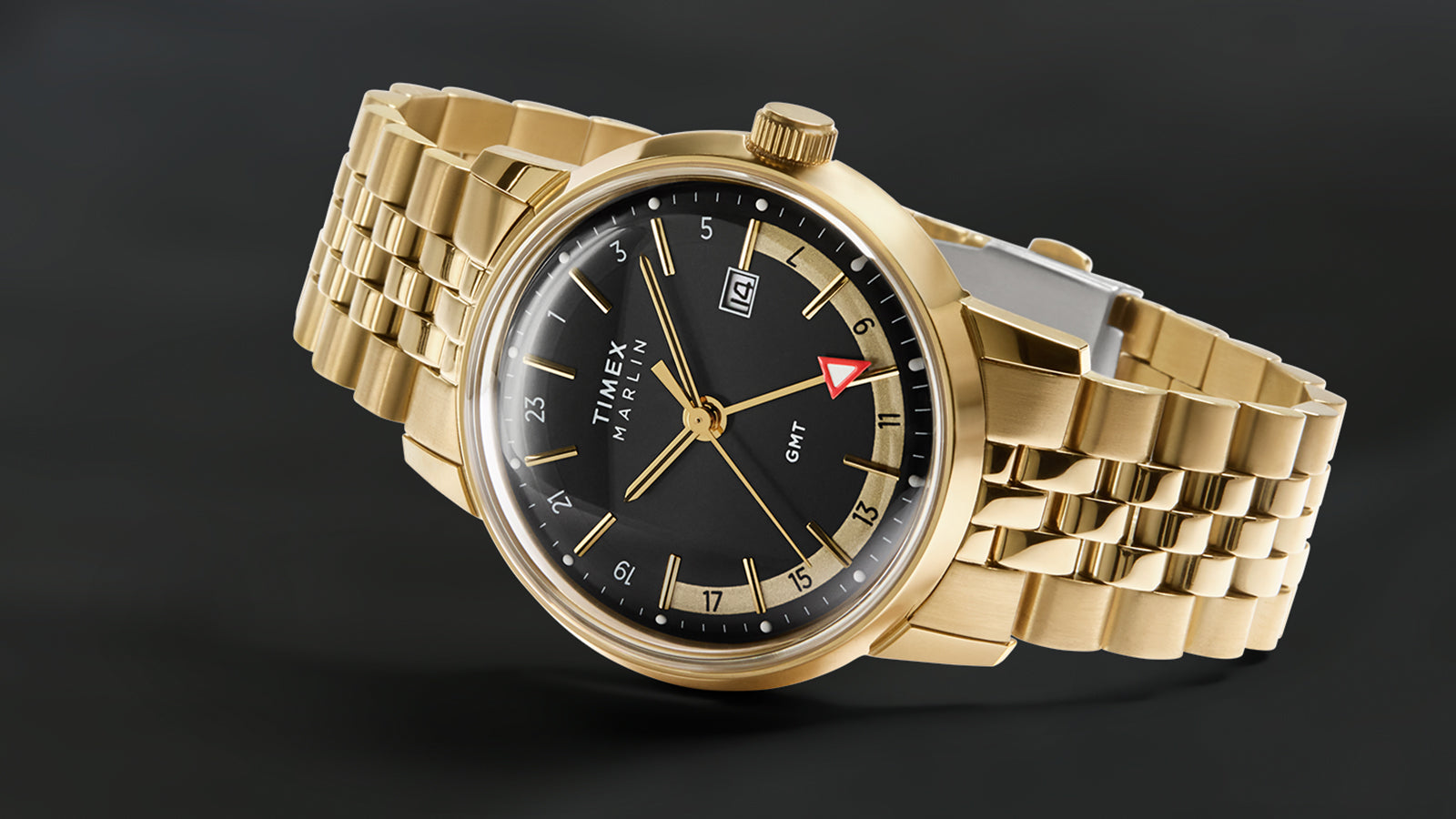 A studio shot of the Timex Marlin® Quartz GMT angled to reveal the brushed gold-tone bracelet and case. The watch face features a black dial with gold-tone hour markers, date window, and the distinctive red GMT hand.