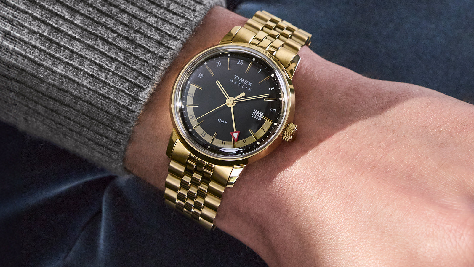 A close-up of a person wearing the Timex Marlin® Quartz GMT with a gold-tone case and bracelet. The watch features a black dial, gold-tone hands and indices, a red GMT hand, and a date display at 3 o'clock.