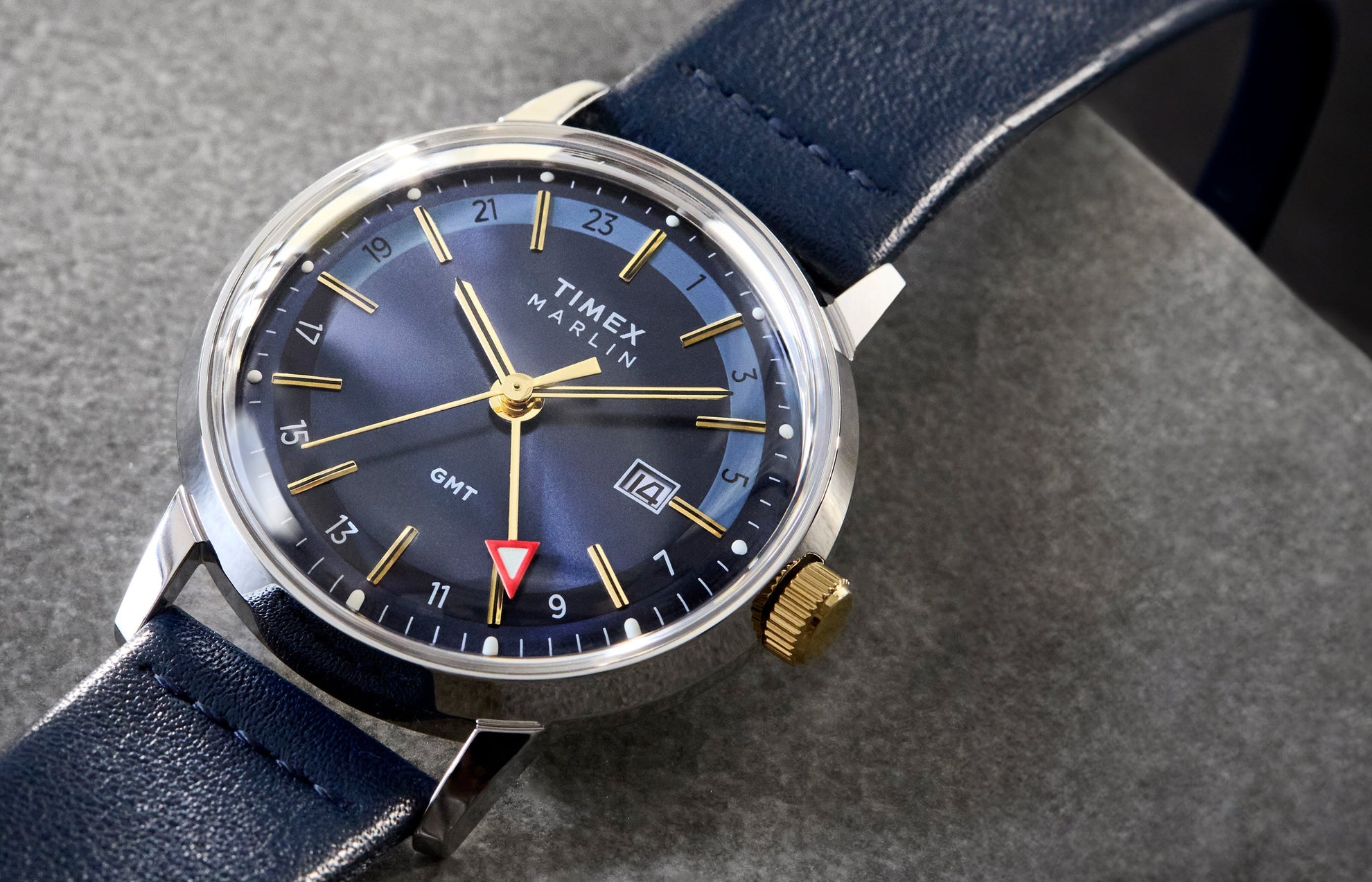 The Timex Marlin® Quartz GMT resting on a stone-textured surface. The rich blue dial, highlighted by golden hour markers, is paired with a polished silver-tone case and navy leather strap.