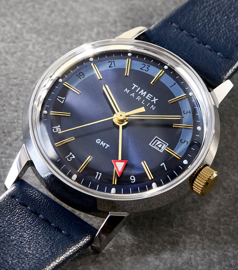 The Timex Marlin® Quartz GMT resting on a stone-textured surface. The rich blue dial, highlighted by golden hour markers, is paired with a polished silver-tone case and navy leather strap.