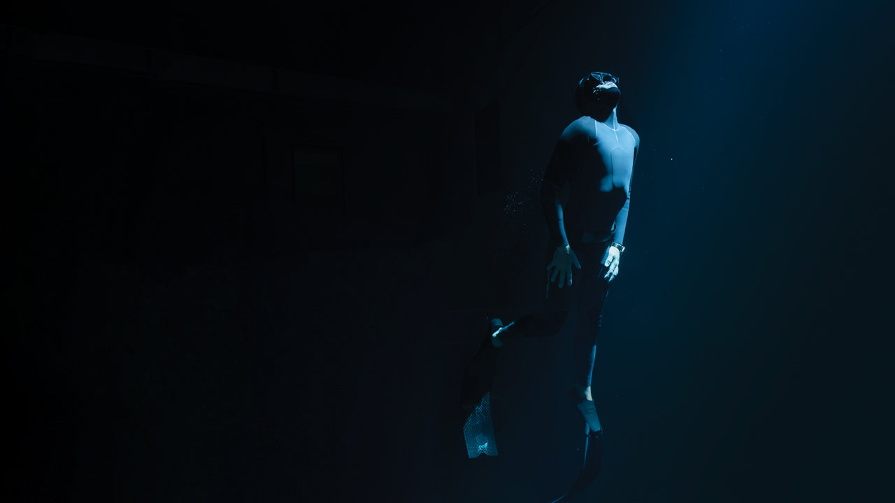 Freediver in a black wetsuit and fins ascends toward the surface in deep water, illuminated by a shaft of light from above.