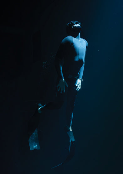 Freediver in a black wetsuit and fins ascends toward the surface in deep water, illuminated by a shaft of light from above.