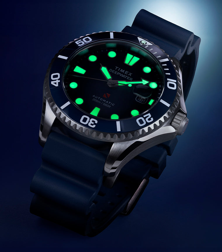 Close-up of the Timex Deepwater Meridian 200 automatic dive watch with luminous markers glowing green against a dark blue background.