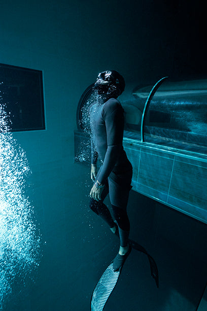 Freediver in a wetsuit illuminated by a beam of light underwater, wearing the Timex Deepwater Meridian 200 dive watch while floating upward in the deep.