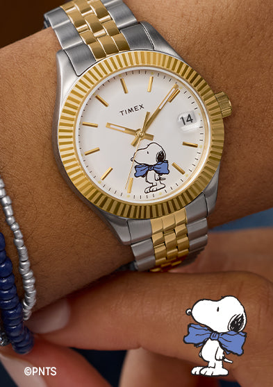 Timex x Peanuts® Timex Legacy watch on wrist with silver and gold-tone bracelet, gold fluted bezel, white dial featuring Snoopy wearing a blue bow tie at 6 o’clock, date window at 3 o’clock, paired with silver and navy beaded bracelets.